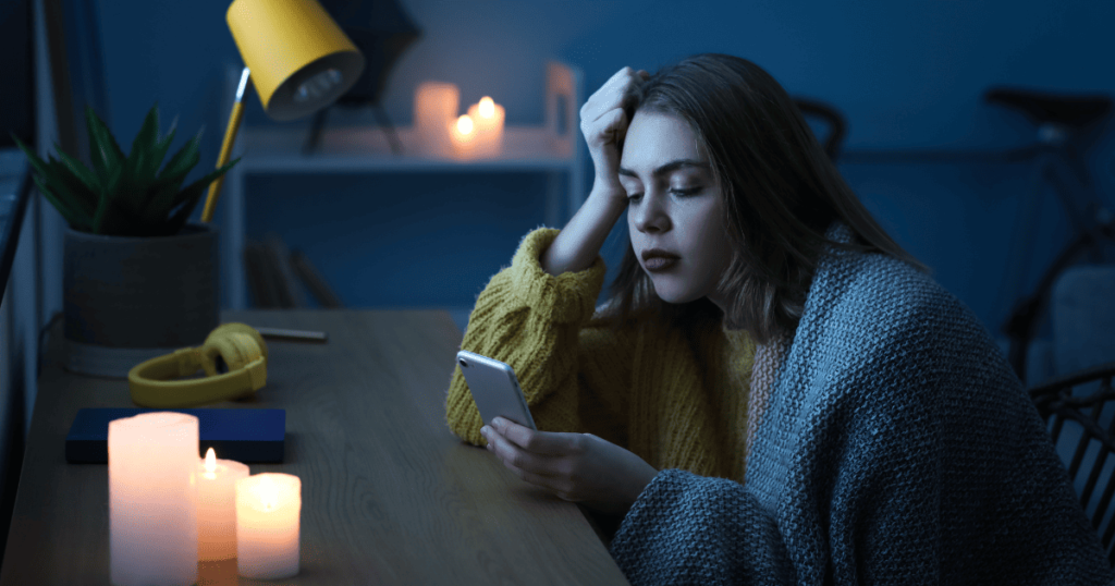 An image of a woman sitting at a desk surrounded by lit candles looking with despair at her mobile phone because her home is in a blackout and has no electricity