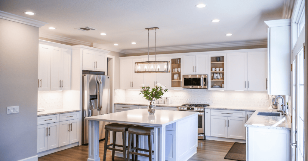 Image of a clean white kitchen with an over the island hanging light fixture, undercabinet lighting, and ceiling mounted spotlights