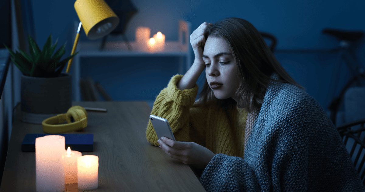 An image of a woman sitting at a desk surrounded by lit candles looking with despair at her mobile phone because her home is in a blackout and has no electricity