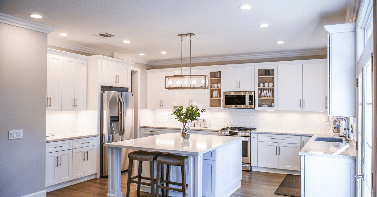 Image of a clean white kitchen with an over the island hanging light fixture, undercabinet lighting, and ceiling mounted spotlights